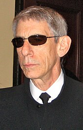 An unsmiling gray-haired man wearing sunglasses and a black shirt with a white collar looks off-screen.