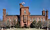 A brick building, reminiscent of a castle, slightly obscured by bare trees. There is a large central tower with the entrance at its base as well as smaller tower at each corner of the building, each with a varying design.