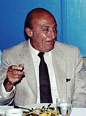 A photograph of an elderly man, seated and wearing a light-gray suit and tie.