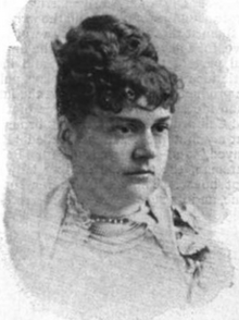 Portrait of a white woman with dark curly hair in an updo. She is wearing a light-colored garment, with beads at her throat.