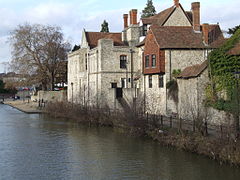 A stone built house with red-tiled roof, overlooking a river. Behind a blue sky with white clouds.