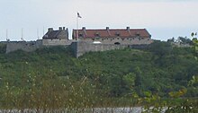 The fort is shown above foreground greenery, photographed through some grass-like plants. Some of the fort's star-points are visible, and a flag is flying from its flagstaff. The sky is a hazy blue-white.