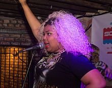 A woman with blonde hair with her right arm raised and her left on her hip sings into a mic in a dark brick room.