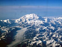 In an aerial image, a mountain is surrounded by many smaller mountains and a glacier.