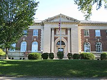 A two-story brick building with pillars, arched windows, and a flagpole in front. A tree is located on the left, and a lawn is in the foreground.