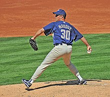 A man in a navy blue baseball jersey prepares to throw a baseball from a pitcher's mound with his right hand. His jersey reads "Maddux" in small tan print and "30" in larger tan print. He is wearing a navy blue baseball cap and gray baseball pants.