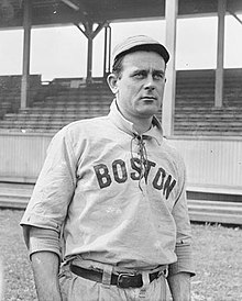 A man in a white cap and baseball jersey with a high collar and "Boston" written across the chest standing in front of a grandstand