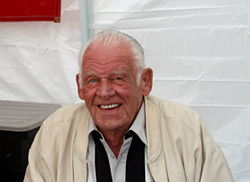 A man wearing a vanilla-colored jacket, with a white collared dress shirt and a black scarf around his neck