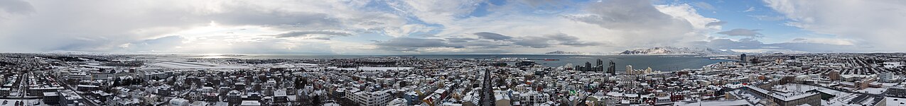 Buildings in the city center of Reykjavik covered in snow, the airport on the left, the harbor in the middle and behind that the Atlantic Ocean, in the distance mountains covered with snow and a partly broken cloud cover.