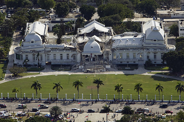 An aerial photograph of a white building with a green lawn and palm trees. Onlooking are standing behind the white fence on the road. The roof appears to have collapsed upon the story beneath and concert rumble is blocking the front steps.