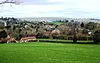 multiple buildings with red and grey roofs nestled amongst trees. Church tower to the left. Foreground is grassy fields and hedgerows. Background is hills.