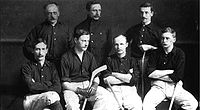 One row of four men seated with three men standing behind. Several have hockey sticks