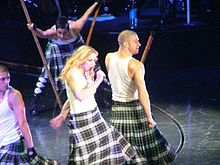 Madonna wearing a white T-shirt and a black-and-white patterned skirt, holding a microphone. She is surrounded by dancers in similar attire, carrying long poles.