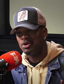 Ne-Yo, who is a man wearing a vest, tie, and hat, standing with his wrist out on a concert stage with people in the background.