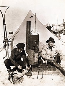 Two men sitting in front of a pointed tent; left-hand man in flat naval cap is skinning a penguin, right-hand man wears a wide-brimmed hat. Between them is a tall stove. Other equipment is visible in the background