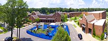 An aerial photograph of the campus featuring a driveway, blue-and-green playground, and several brick buildings. Behind the buildings, only large green trees can be seen.