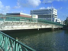 A concrete bridge passing over a body of water is seen from one side. It has green metal decorative railings.