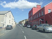 A road between two buildings, one red the other pebbledash, with cars parked on either side.