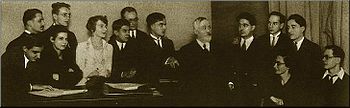 A group of ten young men and three young women, in early 20th-century dress, surround an elderly man with greying hair and beard. On the right some of the group lean over a table with open musical scores.