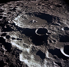 A gray, many-ridged surface from high above. The largest feature is a circular ringed structure with high walled sides and a lower central peak: the entire surface out to the horizon is filled with similar structures that are smaller and overlapping.