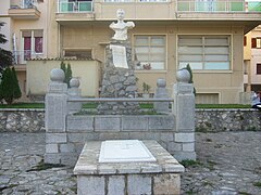 Photo of a tomb of hewn stone, with a marble bust at the back, and an apartment building in the background