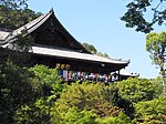 Wooden building with a large hip-and-gable roof built on a hillside. In front of the building there is a wooden railed platform.