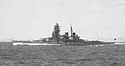 Side view of a dark gray warship at speed. She has two funnels, one mast and a massive forward superstructure.