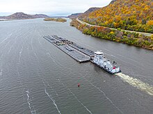 A small boat pushes a large flat barge down a wide river in the fall