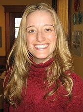 A young caucasian woman with blond hair and a red turtleneck sweater smiles at the camera