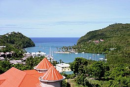 A bay is surrounded by vegetation-covered hills and filled with white sailboats. On the land are houses and building, the closer ones with red roofs and the further ones with white roofs. The entrance to the bay is in the distance, where the water begins to turn a darker blue.