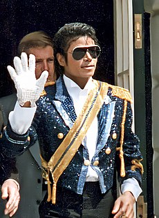 Jackson is wearing sun glasses and a black sequinned jacket with gold shoulders and a golden sash running diagonally across his chest. His right hand is raised in a wave; he is wearing a white glove on it. His skin is dark