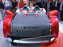 A matte black and bright red two-seat roadster from the front, indoors on a platform with a crowd looking on behind.