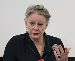 Deirdre McCloskey wearing a dark striped suit top, gesticulating with left hand, appearing to speak, while looking right of camera