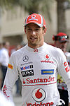 Jenson Button wearing a red baseball cap and white racing overalls with sponsors logos