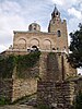 =View of a medieval Byzantine-style church's front facade and bell tower slightly from below, with partially destroyed stone walls in the foreground