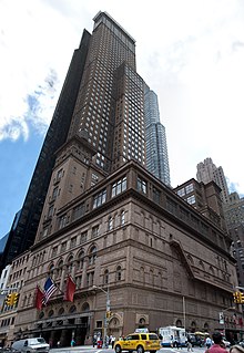 Image of a brown building, taken from across a street intersection. In the foreground are cars and pedestrians; in the background are other buildings and a cloudy sky.