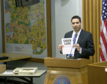 Man holds up a paper labeled "ender card" next to a machine that processes ballots and tabulates results.