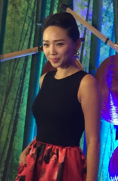 A woman wearing a red floral dress with a black top, standing against a backdrop that has bamboos attached with several Asian conical hats and wood sticks.