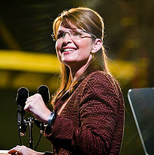 A woman with glasses and long brown hair looks up, and to her left slightly, while speaking at a podium into a microphone, in front of an audience.