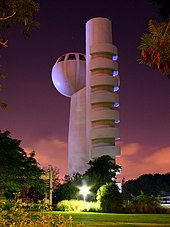 A night-time image of a gray windowless tower, with an egg-shaped windowed observation deck on top. Next to it is a low building, grass, and many trees and bushes.