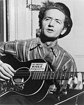 Half-length photo of a middle-aged man sitting in front of a closed door and playing a guitar and singing. His wavy black hair is partly covered by a black hat tipped at a rakish angle. He wears a striped flannel work shirt. His black guitar has a sign on it that says, "This machine kills fascists".