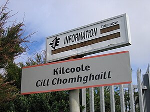 The name of the station i.e. Kilcoole, Cill Chomhghaill written in black font on white board with orange border on top and bottom. Another board with "Information" is written on white board above the name of the station. The board provides current time, and details of next train i.e. name and expected time of arrival. The details at the time of the picture are not clear. The picture is taken during the day.