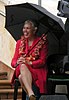 A Samoan lady wearing a red dress and flower lei, sitting on a bench with an umbrella