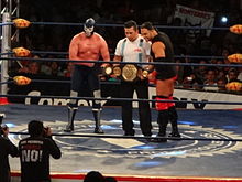 El Texano Jr. and Blue Demon Jr. holding the AAA Mega Championship belt in a wrestling ring.