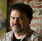 A portrait of a man with curly black hair and a salt-and-pepper beard facing the left of the screen, eyes turned towards the viewer