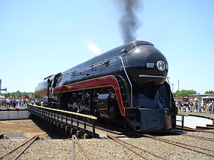 A black streamlined steam locomotive with a 4-8-4 wheel arrangement (four leading wheels, eight driving wheels, and four trailing wheels) and its tender