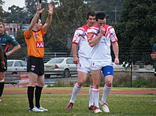 A male referee in an orange shirt signals to two male players in white and red uniforms. The referee stands with his hands up and fingers spread.