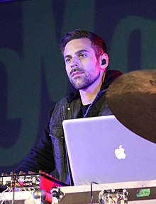 A Caucasian male with earbuds and a black leather jacket staring at an off screen entity with a MacBook laptop in the foreground