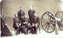 A photograph depicting a group of five uniformed men posed between a pyramid of artillery shells on the left and a wheeled field artillery piece on the right