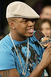 A man wearing a printed blue t-shirt, silver jewelry and a cap; he is holding a microphone.